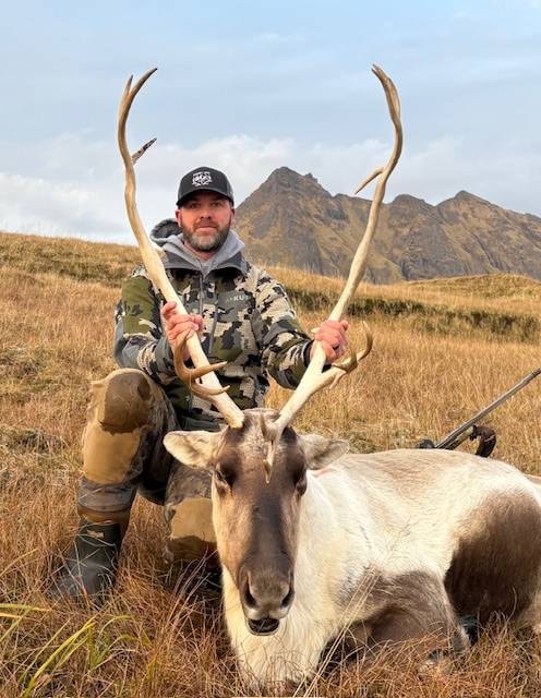 Hunter kneeling with a big Caribou bull, dramatic Adak mountain peak behind