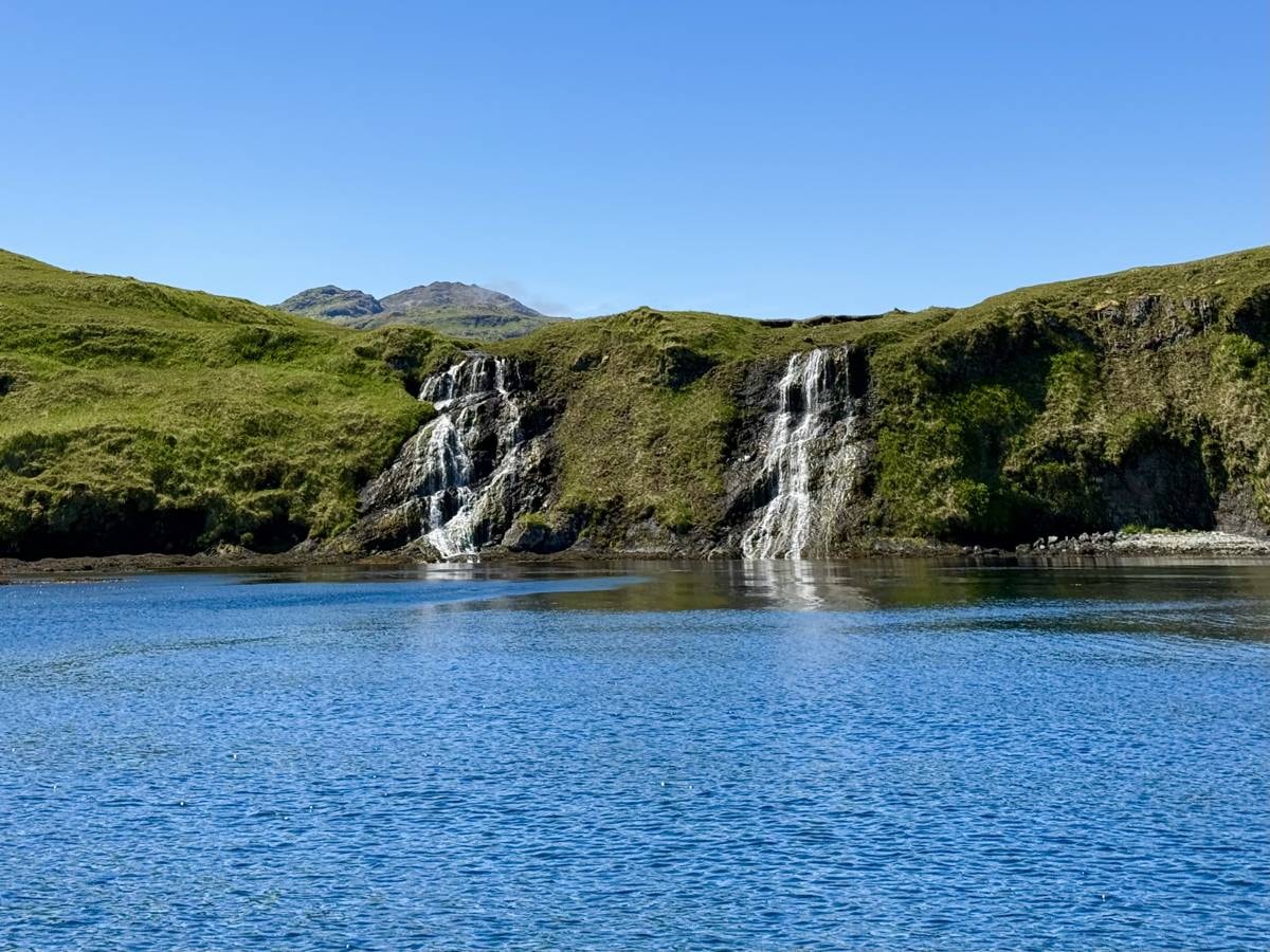 Adak boat on glassy water with twin waterfalls and blue sky
