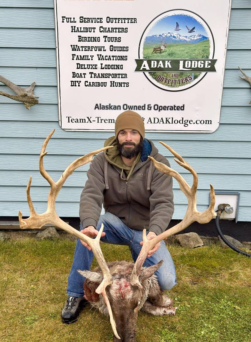 Hunter kneeling with massive Caribou rack at the Adak Lodge sign