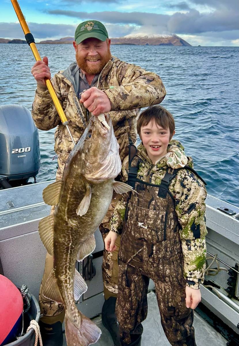 Guide and young boy holding a big Lingcod on the boat, snow-capped Adak island behind — families welcome