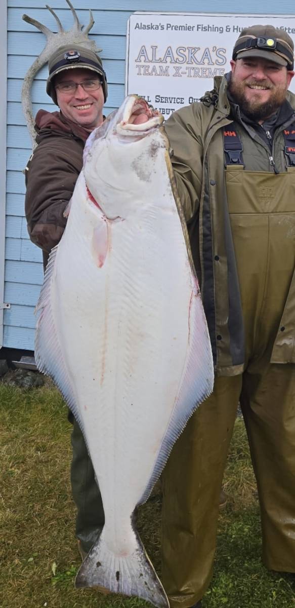 Two anglers with a massive Halibut at the Adak Lodge sign