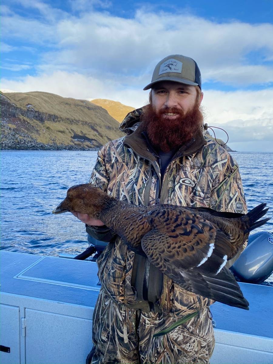Bearded hunter holding a Sea Duck with Aleutian cliffs behind