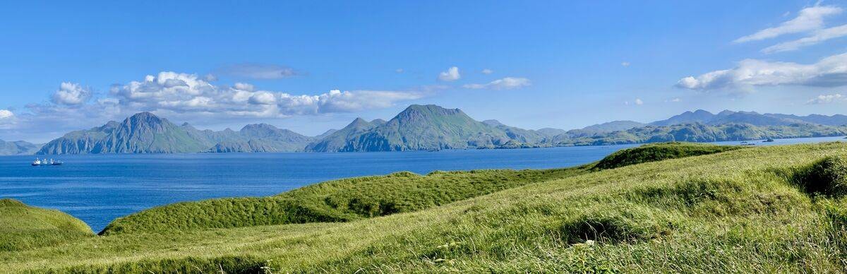Wide panoramic view of Adak Island — rolling green tundra, deep blue ocean, and distant mountain islands