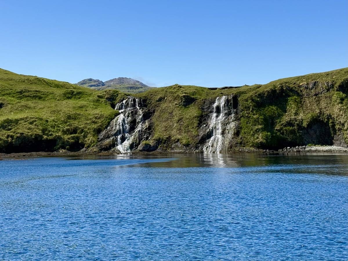 Twin waterfalls cascading into a deep blue Adak cove, green tundra cliffs and clear sky