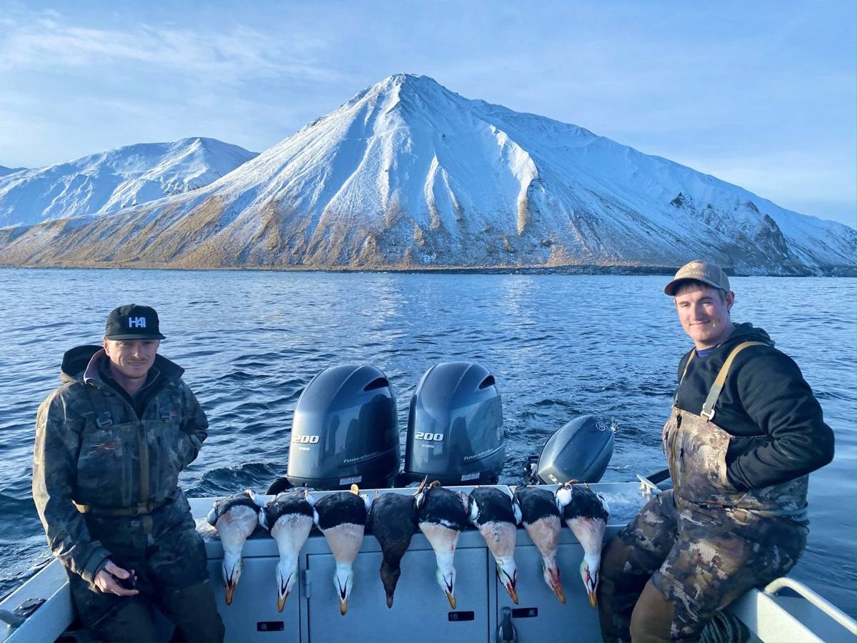 Two hunters on the boat with King Eiders lined up, snow-capped Adak volcano behind